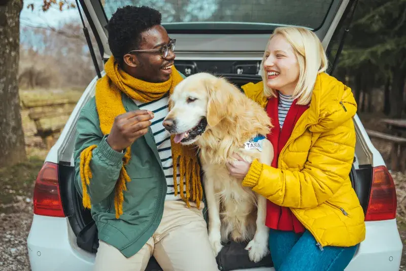 Two people in colorful jackets sit in the open trunk of a parked car with a golden retriever, spending time together at a wooded outdoor location.