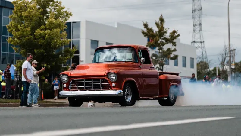 Vintage orange pickup truck driving past spectators at a car show.