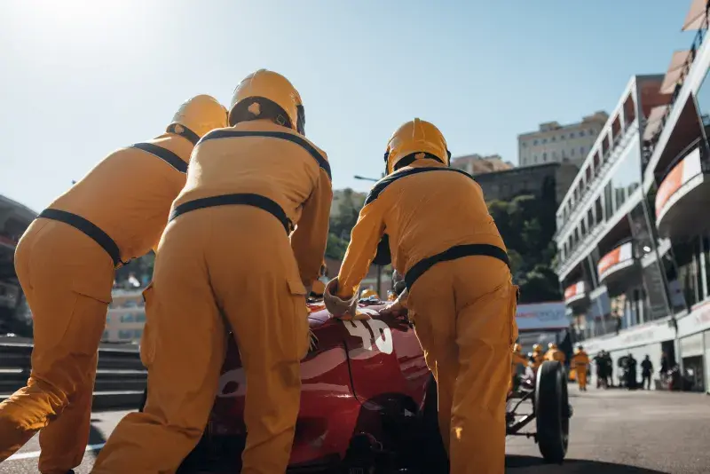 Pit crew members in yellow uniforms pushing a red race car along a racetrack during a motorsport event, with grandstands and buildings visible in the background.