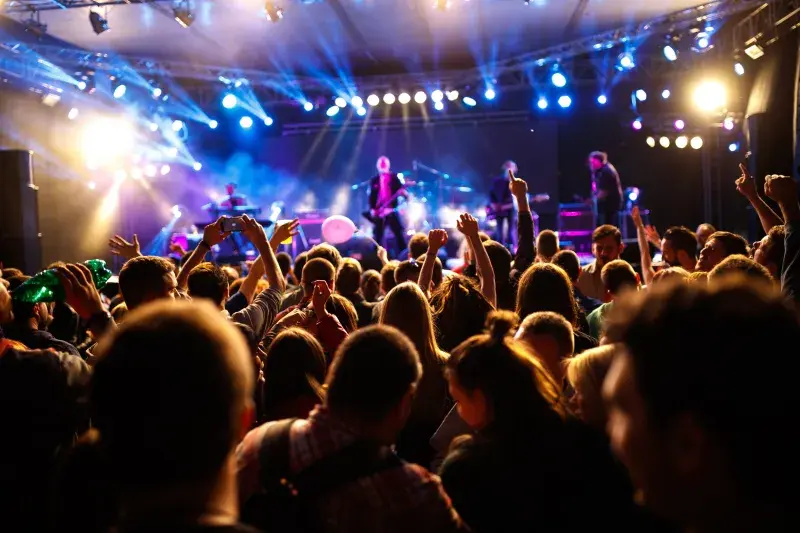 Close-up of a crowd at a live event, with audience members standing shoulder to shoulder and raising their hands toward the stage under warm lighting.