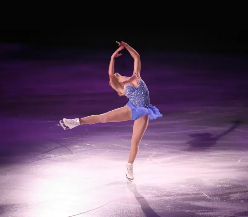 Figure skater performing on an ice rink under purple stage lighting, balancing on one skate with arms extended overhead during a solo routine.