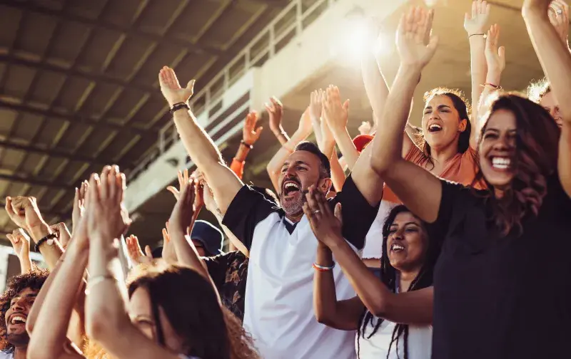 Audience members in stadium seating raising their hands and cheering during a live sporting or entertainment event in a sunlit venue.