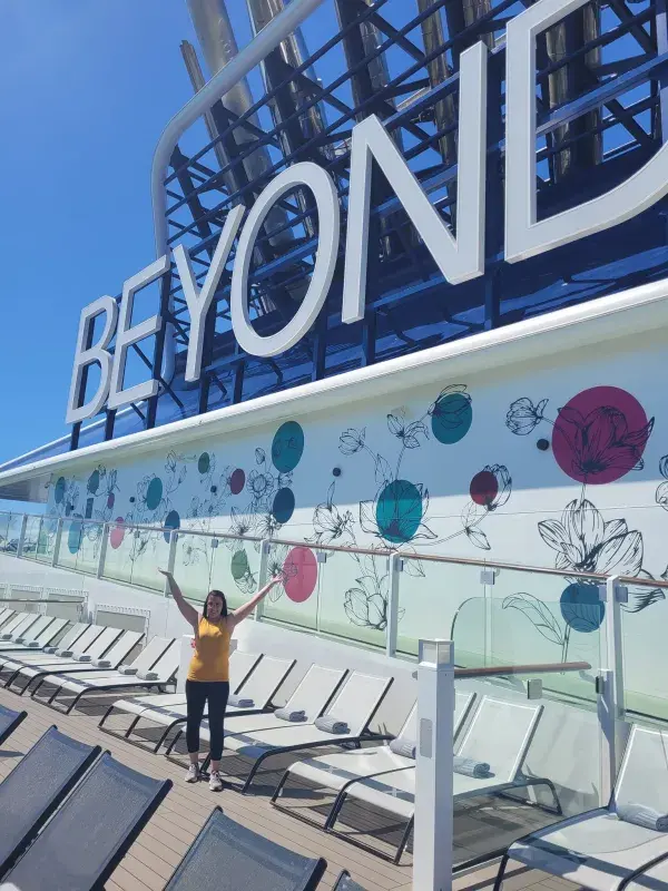 Brittany standing on a cruise ship deck beneath a large “BEYOND” sign, surrounded by lounge chairs and decorative floral artwork.