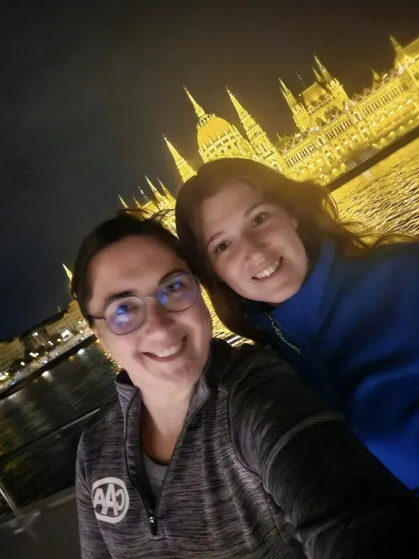 Two travellers posing along the Danube River at night with the illuminated Hungarian Parliament Building in Budapest in the background.