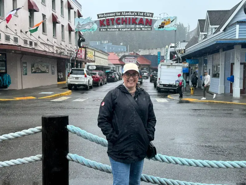 Brittany standing beneath the Welcome to Ketchikan sign on a rainy day in Alaska's historic downtown waterfront district.