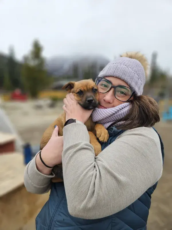 Brittany wearing a winter hat and glasses holding a small brown puppy outdoors, with a blurred mountain landscape in the background.