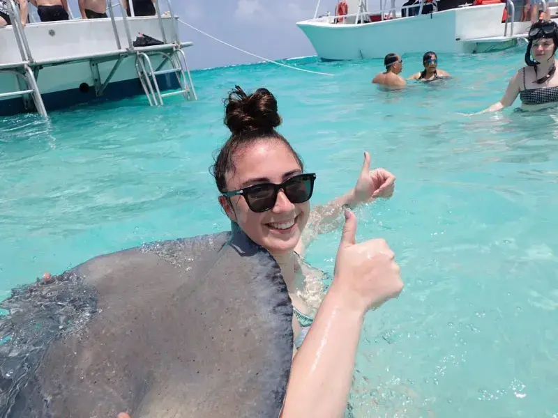 Brittany wearing sunglasses in clear turquoise water giving a thumbs up while a stingray swims nearby, with boats in the background.